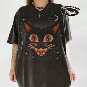 May include: A charcoal grey short-sleeve t-shirt featuring a large, stylised graphic of a cat face with orange eyes and a textured, knit-like pattern. The design includes small white stars and a crescent moon. A label reads COMFORT COLORS Pepper.