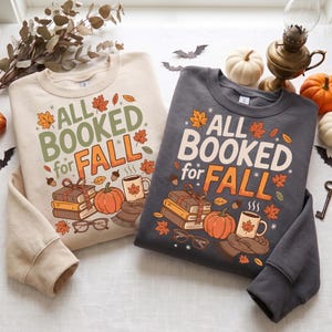 May include: Two crewneck sweatshirts, one cream and one dark gray, each featuring a graphic print that reads ALL BOOKED for FALL. The design includes a stack of books tied with a ribbon, a pumpkin, a steaming mug with a leaf, and reading glasses.
