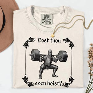 Op de afbeelding: Cr&egrave;mekleurig T-shirt met een zwarte afbeelding van een ridder die gewichten heft, omlijst door sierlijke krullen. De tekst "Dost thou even hoist?" staat boven en onder de ridder. Een gouden ketting met een sterhanger ligt op het shirt.