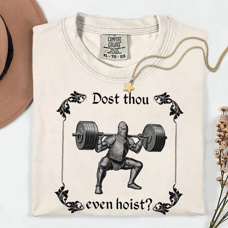 Op de afbeelding: Cr&egrave;mekleurig T-shirt met een zwarte afbeelding van een ridder die gewichten heft, omlijst door sierlijke krullen. De tekst "Dost thou even hoist?" staat boven en onder de ridder. Een gouden ketting met een sterhanger ligt op het shirt.