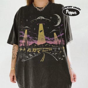 May include: A charcoal gray t-shirt featuring a graphic of three UFOs hovering over a desert landscape with cacti, a road, and a sign that reads ROSWELL NM with an alien icon. The design includes yellow light beams, a crescent moon, and stars.