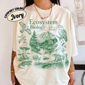 May include: An ivory t-shirt featuring a green graphic print with the text Ecosystem Biology Life in Balance. The design includes illustrations of a mountain landscape, a forest, a bird, an owl, mushrooms, a snail, a bee, a frog, and a lizard.