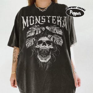 May include: A charcoal gray short-sleeve t-shirt featuring a white graphic print of a skull with monstera leaves emerging from the top. The text MONSTERA is printed in a distressed font above the skull. A circular badge reads COMFORT COLORS Pepper.