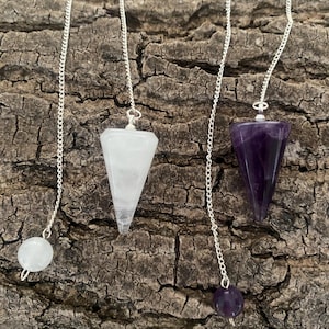 May include: Two dowsing pendulums on silver-toned chains. One pendulum is a white, translucent, cone-shaped stone with a matching round bead at the end of the chain. The other is a deep purple, cone-shaped amethyst stone with a matching purple bead.