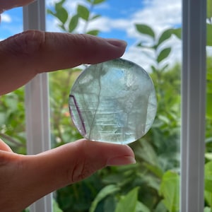May include: A circular, polished fluorite stone featuring translucent pale green and clear sections with a distinct, thin purple band running along one side. The mineral displays natural internal fractures and variations in colour throughout its surface.