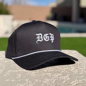 May include: Black baseball cap with a flat brim and white rope detail. The cap features the embroidered letters "DGP" in a gothic font. The hat is sitting on a stone surface, with a blurred green background.