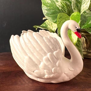 May include: A white ceramic swan planter with a red beak and black accents. The swan has detailed feather textures and is positioned on a wooden surface. The planter is designed to hold small plants or flowers.