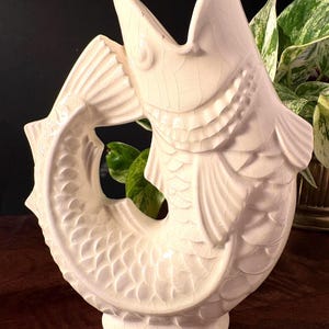 May include: A cream ceramic fish sculpture with an open mouth and curved tail. The fish features detailed scales and fins, and a crackled glaze finish. This decorative item is a unique piece.