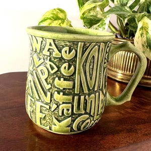 May include: Green ceramic coffee mug with embossed lettering. The mug features the words "WA, Kaffee, Kaffe, Coffee, UNL" in a repeating pattern. The handle has a unique, textured design. The mug is a light green color.
