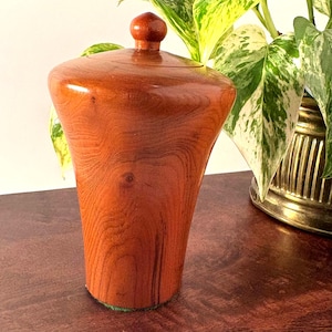 May include: A handcrafted wooden urn with a lid. The urn is a warm reddish-brown color with visible wood grain. The lid has a small, rounded knob. The urn is displayed on a dark wooden surface, with a green base.