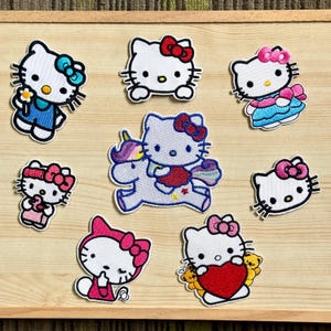 Op de afbeelding: Een verzameling geborduurde patches met het iconische Hello Kitty-personage in verschillende poses en outfits. De patches tonen Hello Kitty met strikken, bloemen en een hart, met een eenhoorn en in een jurk. De patches zijn wit, roze, rood, blauw en geel.