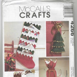 May include: A sewing pattern for making Christmas stockings and a wine bottle holder. The pattern includes four different designs: a red stocking with white trim and gold bells, a brown stocking with a diamond pattern, a green stocking with white trim and gold bells, and a red and green wine bottle holder with a gold bow.