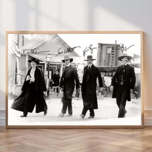 Puede incluir: Impresión enmarcada en blanco y negro que presenta a cuatro hombres con atuendo de época caminando por una calle polvorienta. La imagen tiene firmas encima de los hombres. Los hombres llevan sombreros y abrigos largos, evocando un tema del Oeste.
