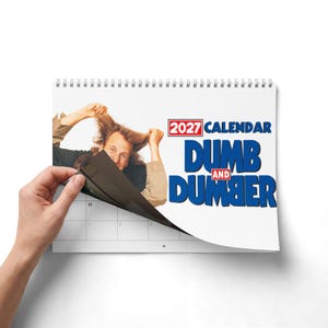 Może przedstawiać: Kalendarz ścienny 2027 Dumb and Dumber z białą spiralą. Okładka zawiera zdjęcie osoby z potarganymi włosami oraz napis 2027 CALENDAR DUMB AND DUMBER w kolorze niebieskim i czerwonym. Kalendarz jest częściowo otwarty, ukazując stronę z siatką.