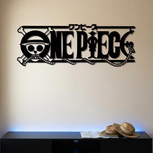 Può includere: Un'opera d'arte da parete in metallo nero che raffigura il logo di One Piece con l'iconico emblema del teschio con ossa incrociate a sinistra, il testo "ONE PIECE" in grassetto maiuscolo e caratteri giapponesi sopra il titolo, collegati da una corda.