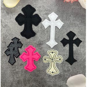 Puede incluir: Un conjunto de parches decorativos con forma de cruz en varios colores, incluyendo negro, blanco, gris y rosa. Los parches tienen costuras detalladas y están dispuestos sobre una superficie gris. Adecuado para ropa o proyectos de manualidades.