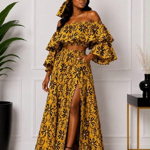 May include: A two-piece set featuring a mustard yellow off-the-shoulder crop top with ruffled sleeves and a matching high-waisted maxi skirt with a front slit. Both pieces feature a dark brown floral and geometric pattern and a head wrap.