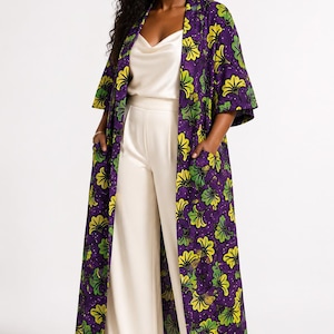 May include: A long, open-front duster coat in a deep purple fabric featuring a repeating pattern of yellow and green floral motifs. The coat has three-quarter length sleeves and two front patch pockets, worn over a cream-colored camisole and trousers.