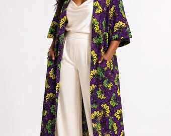 May include: A long, open-front duster coat in a deep purple fabric featuring a repeating pattern of yellow and green floral motifs. The coat has three-quarter length sleeves and two front patch pockets, worn over a cream-colored camisole and trousers.