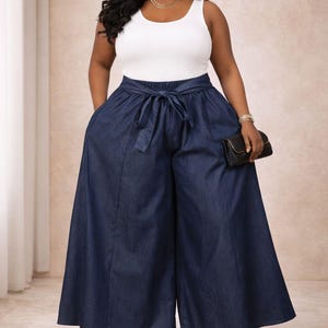 K&ouml;nnte beinhalten: Eine Frau tr&auml;gt ein wei&szlig;es Tanktop und eine dunkelblaue Jeanshose mit weitem Bein. Die Hose hat einen Bindeg&uuml;rtel in der Taille und Taschen. Sie tr&auml;gt au&szlig;erdem Goldschmuck und h&auml;lt eine schwarze Clutch.