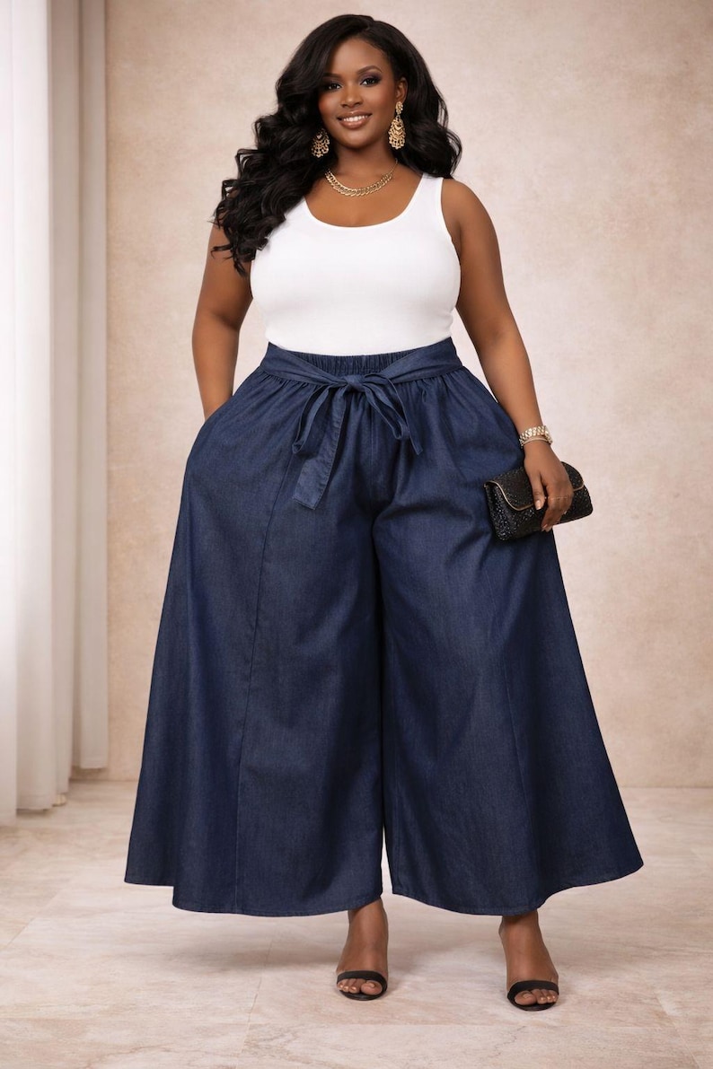 K&ouml;nnte beinhalten: Eine Frau tr&auml;gt ein wei&szlig;es Tanktop und eine dunkelblaue Jeanshose mit weitem Bein. Die Hose hat einen Bindeg&uuml;rtel in der Taille und Taschen. Sie tr&auml;gt au&szlig;erdem Goldschmuck und h&auml;lt eine schwarze Clutch.