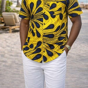 May include: A short-sleeved button-down shirt with a yellow background and a blue teardrop pattern. The shirt is paired with white pants. A gold watch is visible on the wrist.
