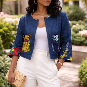 May include: A denim jacket with colorful patterned accents on the sleeves and pockets. The jacket is paired with white pants and a straw clutch. The model is wearing sunglasses and a straw hat.