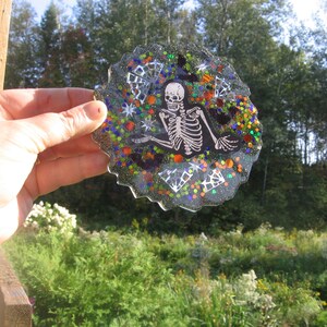 May include: A round, black, glitter-covered glass ornament with a white skeleton design. The skeleton is sitting with its legs crossed and its arms outstretched. The ornament is decorated with colorful glitter and white stars.