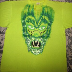 May include: A lime green t-shirt with a green, glitter-covered monster face graphic. The monster has green eyes, a wide mouth with sharp teeth, and gold glitter accents.