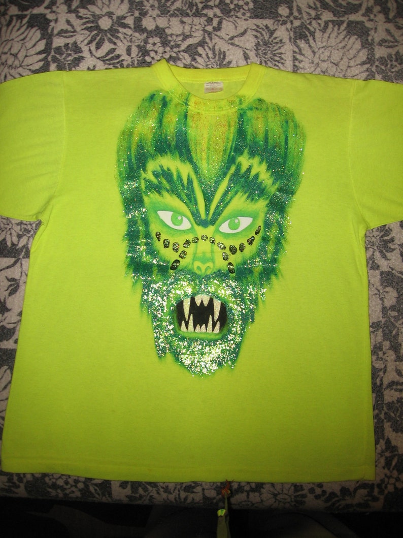 May include: A lime green t-shirt with a green, glitter-covered monster face graphic. The monster has green eyes, a wide mouth with sharp teeth, and gold glitter accents.