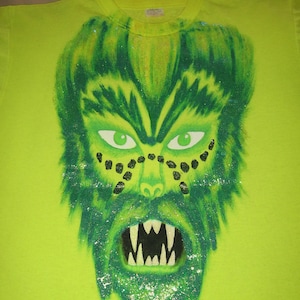 May include: A green t-shirt with a green, glittery monster face printed on the front. The monster has a large mouth with sharp teeth and black eyes.
