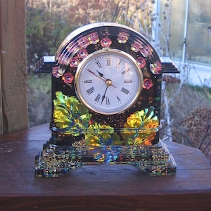 May include: A black and gold clock with a floral design. The clock has Roman numerals and a gold-coloured face. The clock is sitting on a wooden surface.