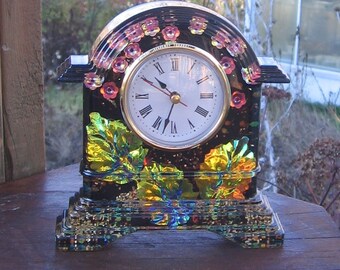 May include: A black and gold clock with a floral design. The clock has Roman numerals and a gold-colored face. The clock is sitting on a wooden surface.