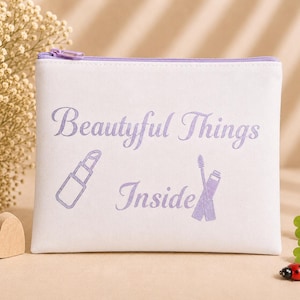 Könnte beinhalten: Eine weiße, rechteckige Reißverschlusstasche mit einem hellvioletten Reißverschluss. Auf der Vorderseite befindet sich der Text "Beautiful Things Inside" in Hellviolett, ergänzt durch hellviolette Strichzeichnungen eines Lippenstifts und einer Wimperntusche.