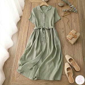 May include: A sage green linen dress with short sleeves, a collared neckline, and a button-down front. The dress features a matching fabric belt tied at the waist and a slit in the skirt. A woven handbag and beige sandals are also visible.
