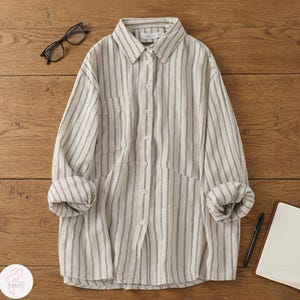 Puede incluir: Camisa a rayas beige y blancas con mangas largas y dos bolsillos delanteros. Las mangas están enrolladas. La camisa tiene la palabra "this" en el cuello. Un par de gafas, un bolígrafo y un bloc de notas están sobre la superficie de madera.
