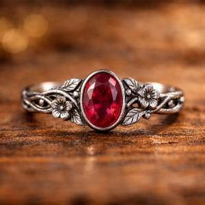 Puede incluir: Un anillo con una piedra preciosa roja de corte ovalado engastada en una banda de plata oxidada. La banda presenta un diseño en relieve detallado de pequeñas flores y hojas que envuelven la piedra central, creando una textura de enredadera.