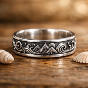 Könnte beinhalten: Ein silberner Ring mit einem detaillierten Berg- und Wellendesign. Der Ring hat ein Band mit einem dunklen, strukturierten Muster aus Bergen, Wellen, Sternen und einer Sonne oder einem Mond. Der Ring wird auf einer Holzoberfläche präsentiert.