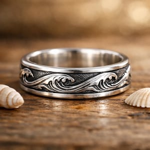 Könnte beinhalten: Ein silberner Ring mit einem Wellenmuster. Der Ring hat ein Band mit einem detaillierten Design von Meereswellen. Der Ring besteht aus einem glänzenden, metallischen Material und befindet sich vor einem hölzernen Hintergrund mit Muscheln.