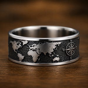 May include: A silver-toned ring with a detailed world map design. The ring features a textured band with continents and a compass rose. The ring is set against a wooden background, highlighting the intricate details of the jewelry.