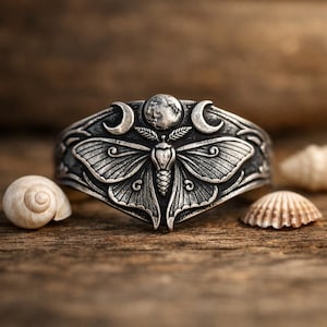 May include: A silver ring with a detailed moth design, crescent moons, and a central sphere. The ring is set against a wooden surface, with seashells in the background. The intricate details and celestial theme make it a unique piece of jewellery.