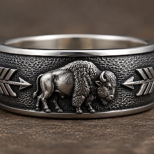 May include: A silver-toned metal ring featuring a detailed relief of a standing bison in the center. The band has a dark, textured background with a polished silver arrow design on either side of the bison, and smooth, polished edges.