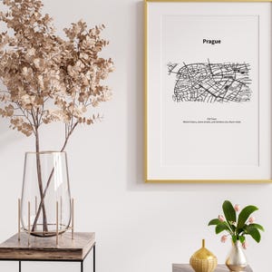 May include: A framed print of a black and white map of Prague, Czech Republic, with the city name above the map. The print is in a gold frame. A clear glass vase with dried branches and two small decorative vases are on a wooden table.