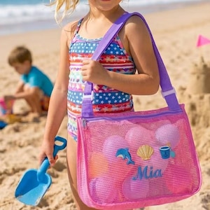 May include: A young person on a sandy beach holds a blue shovel and a pink mesh beach bag. The bag is filled with pink balls and has a purple strap. The bag is decorated with a dolphin, seashell, and a small cup.