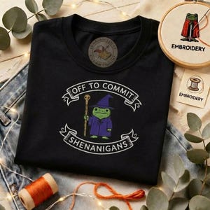 May include: Black t-shirt with an embroidered design featuring a green frog in a wizard's outfit holding a staff. The text "OFF TO COMMIT SHENANIGANS" is arched above and below the frog.