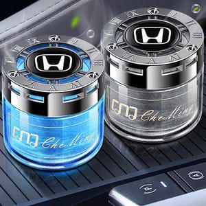 Könnte beinhalten: Zwei Auto-Lufterfrischer mit silbernen Oberteilen und Honda-Logos. Einer enthält blaue Flüssigkeit, der andere graue. Beide haben den Text "Che Ming". Sie stehen auf einer schwarzen Oberfläche, wahrscheinlich einer Autokonsole.