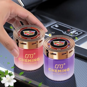 Könnte beinhalten: Zwei Auto-Lufterfrischer, einer rot und einer lila, mit goldfarbenen Deckeln und Rändern. Die Deckel zeigen ein Logo mit einem Wappen-Design. Der Text "CHEMING CAR PERFUME" ist auf der Vorderseite jedes Behälters aufgedruckt. Der Hintergrund ist eine dunkelgraue Oberfläche.