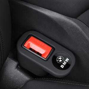 May include: Black seat belt buckle with a red "PRESS" button. The buckle has a black circular emblem with the BMW logo and the word "BMW" in white. The buckle is set against a black leather car seat.