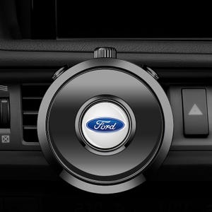 Pode incluir: Suporte de telefone preto para ventilação de carro com design circular. O centro apresenta um círculo branco com o logotipo azul da Ford. O suporte tem um botão na parte superior e dois botões laterais. Fundo do painel do carro.