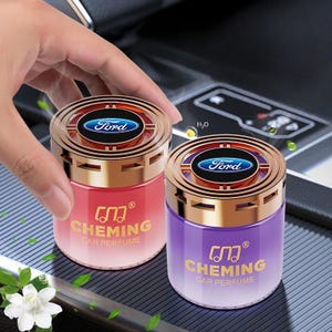 Könnte beinhalten: Zwei Auto-Lufterfrischer mit goldfarbenen Rändern und dem Ford-Logo. Einer ist rot, der andere lila, beide mit dem Text "CHEMING CAR PERFUME" in Gold. Die zylindrischen Erfrischer stehen auf einer dunklen Oberfläche.